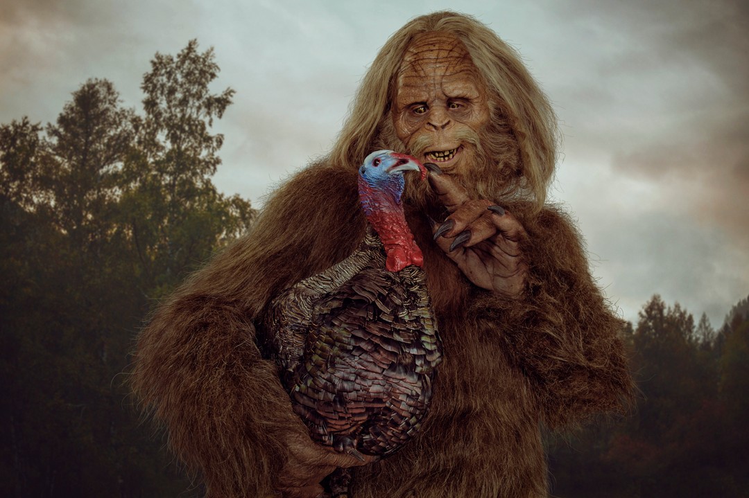 Furry Bigfoot-like character with a brown turkey outdoors against an atmospheric sky and forest background in medium shot.